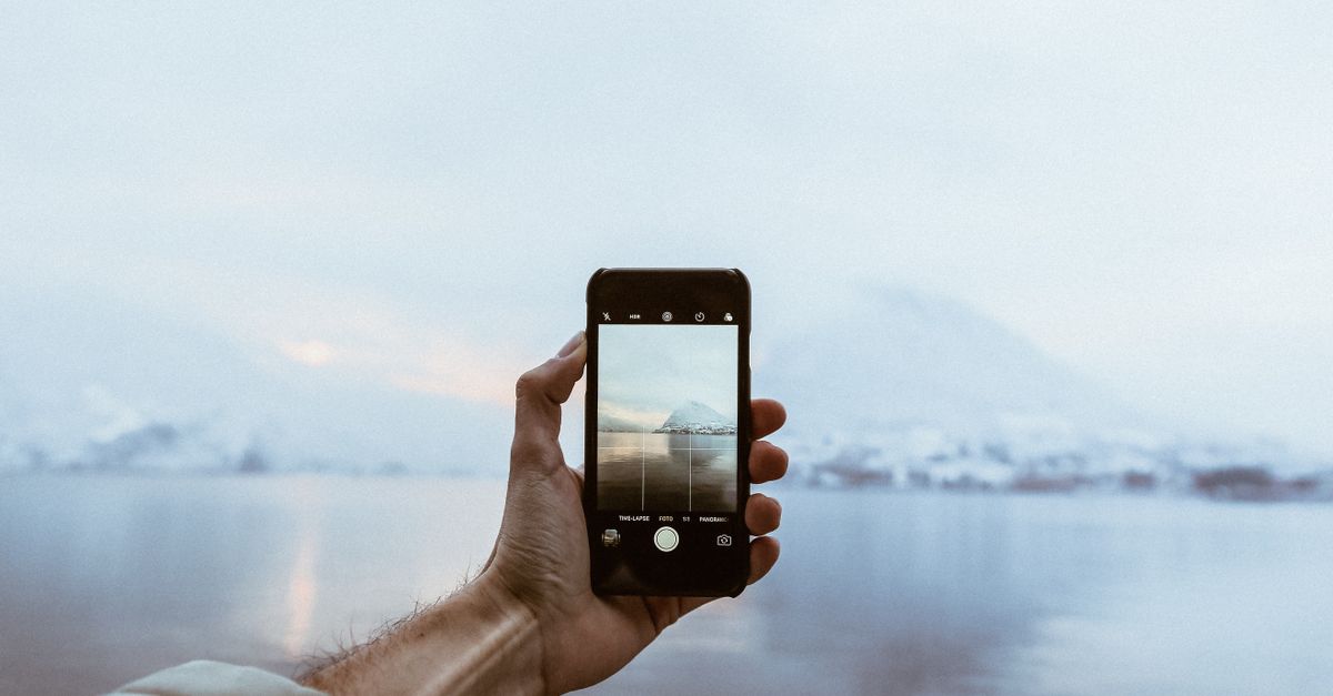 A person holding a smartphone capturing a scenic view of snow-covered mountains by a lake.