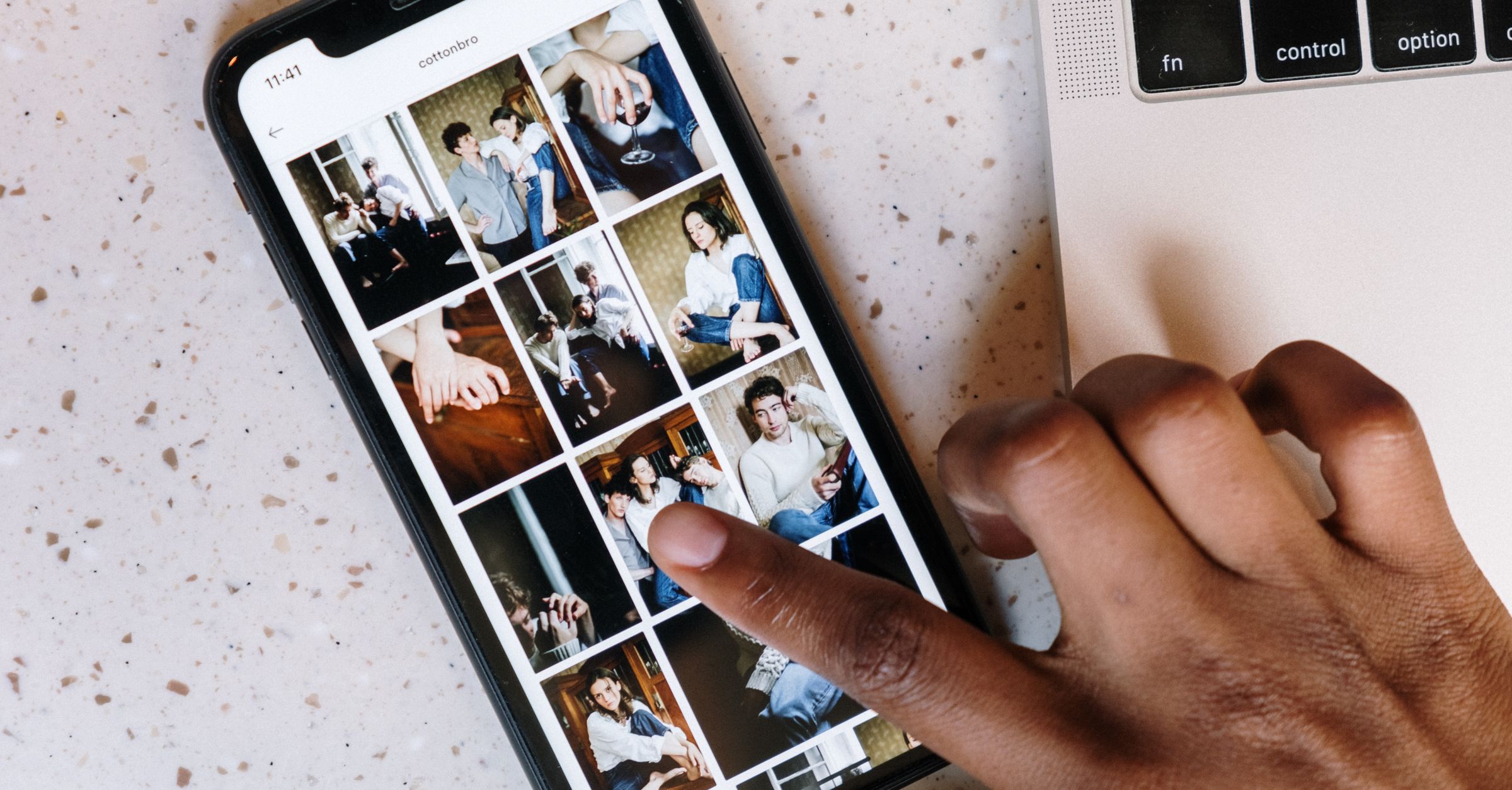 A hand scrolls through a photo gallery on an iPhone beside a laptop.