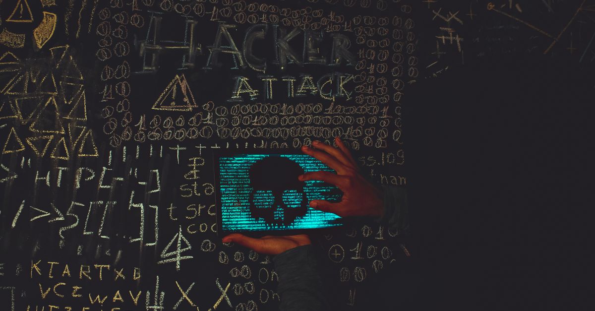 A hacker in a black hoodie using a tablet displaying a skull, surrounded by chalk symbols and 'Hacker Attack' text.
