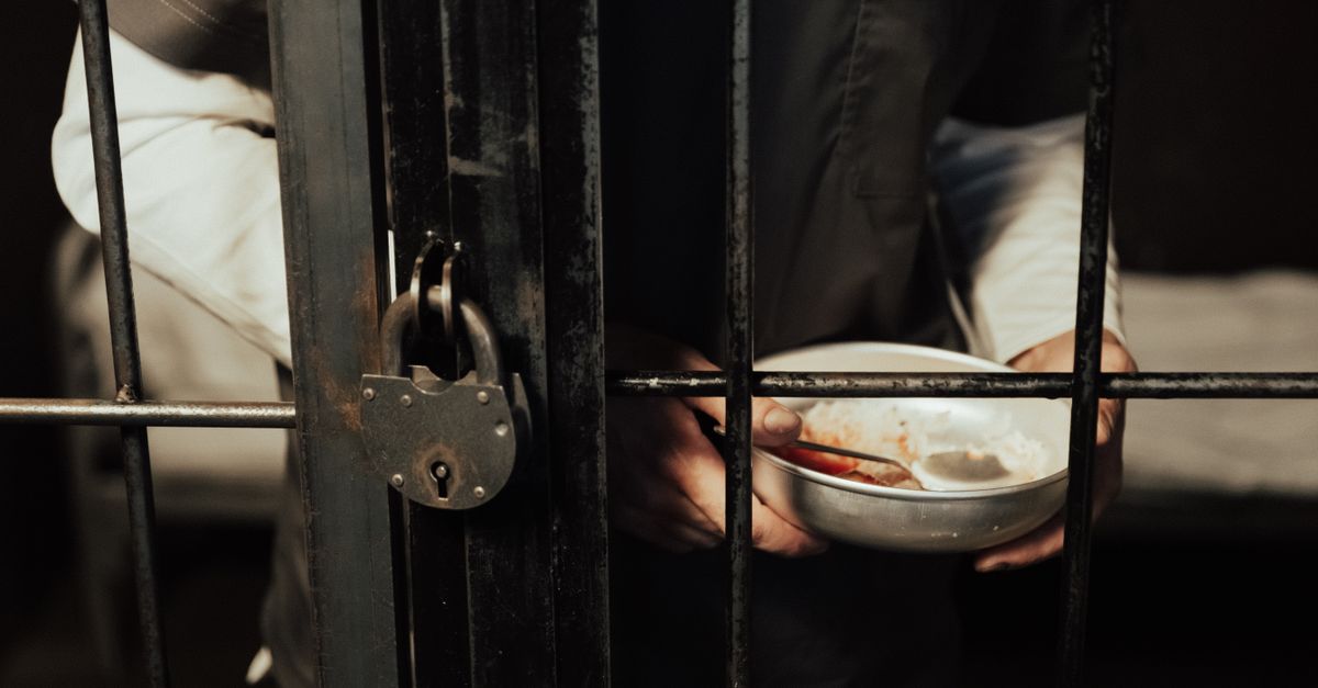 Close-up of a prisoner holding a bowl behind iron bars, illustrating confinement.